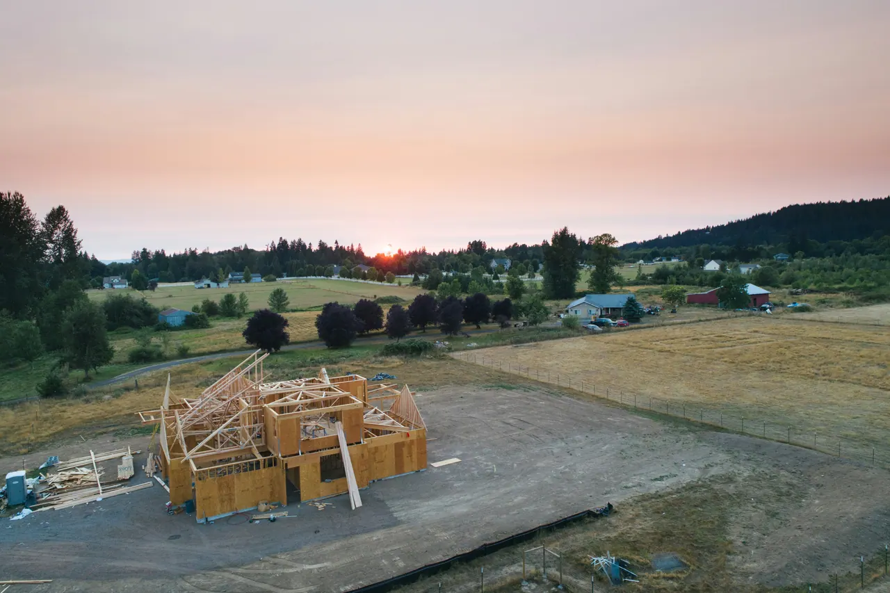 A house being framed on an open rural lot with fields and trees at sunset