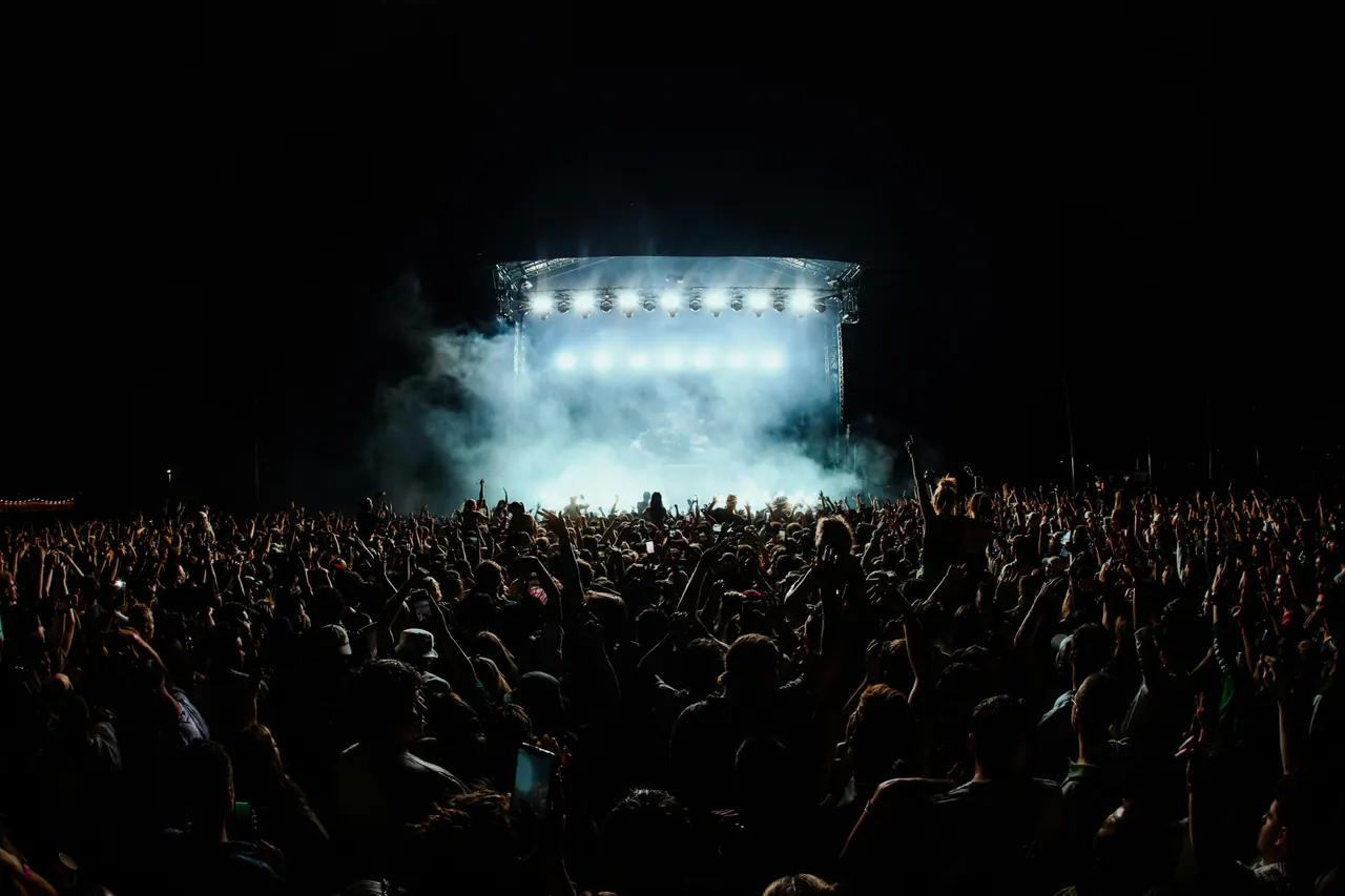 A large crowd in silhouette facing a brightly lit outdoor stage at night