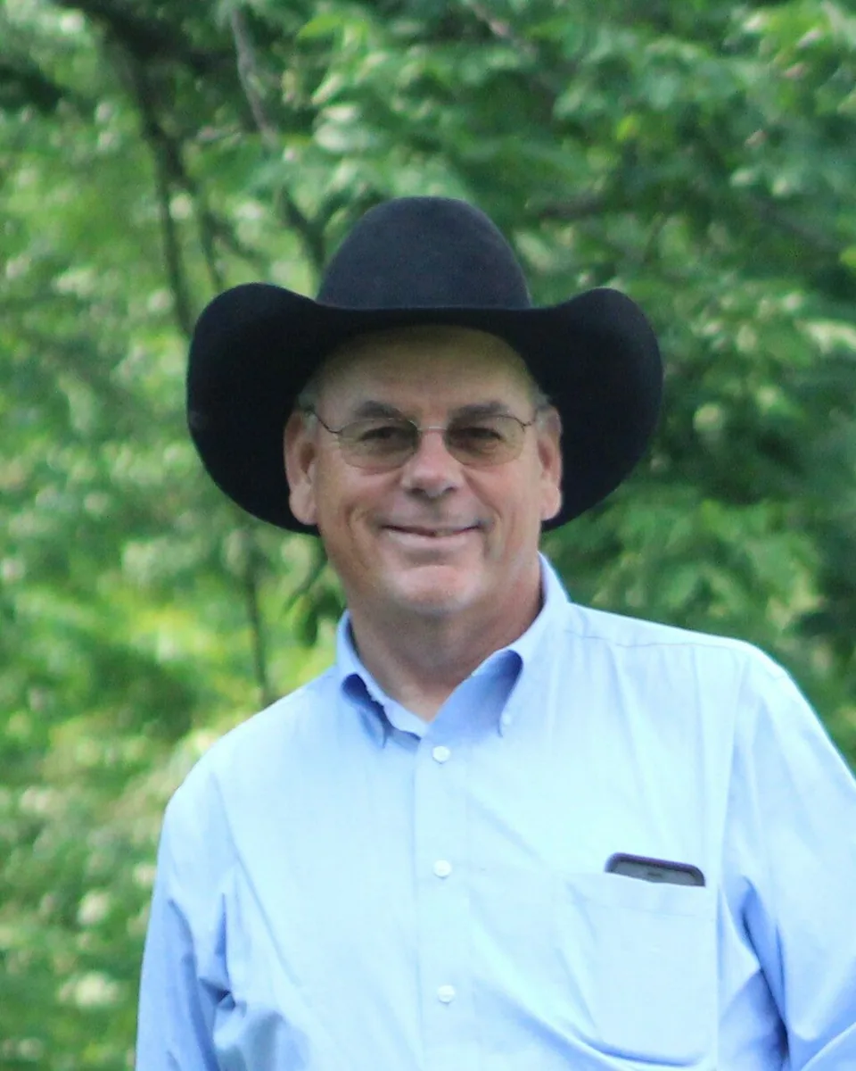 Greg Eliel in a black hat and a blue shirt, smiling under summer trees
